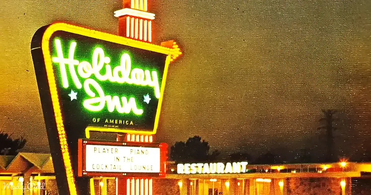 A glowing neon sign for the Holiday Inn of America stands illuminated against a dark sky, with a smaller sign below...