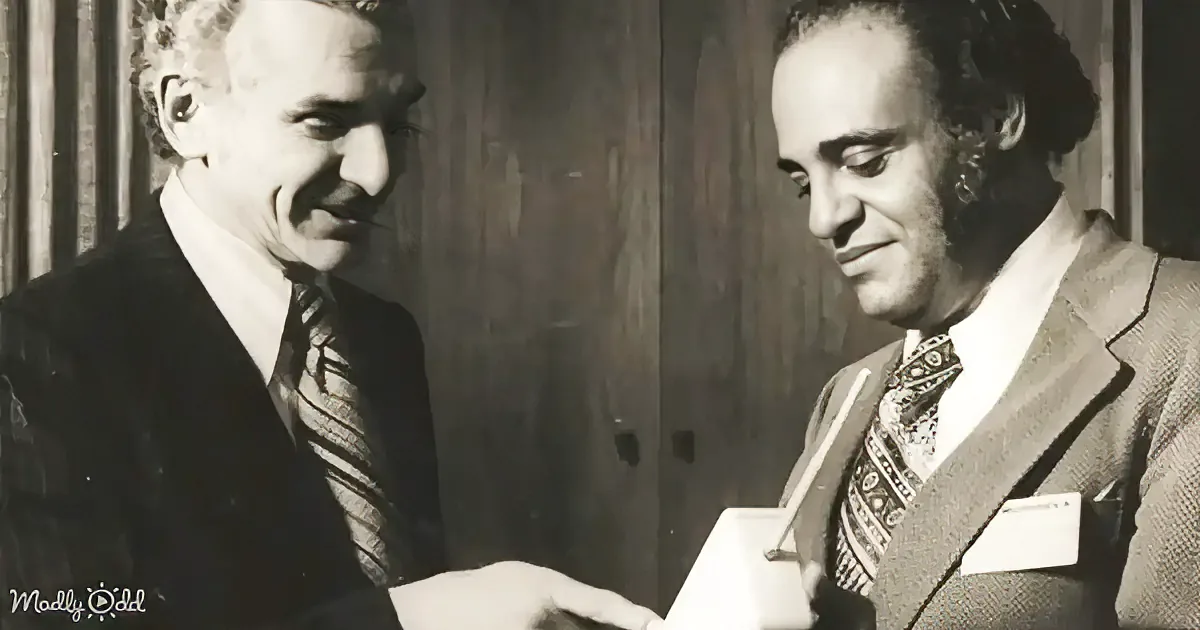 Two men in suits smile, one handing a document to the other, against a wooden backdrop.