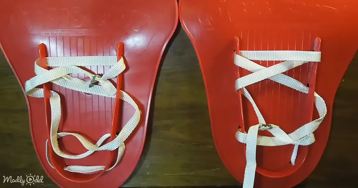 Two red plastic sleds with white straps and metal buckles are shown side by side on a wooden surface.