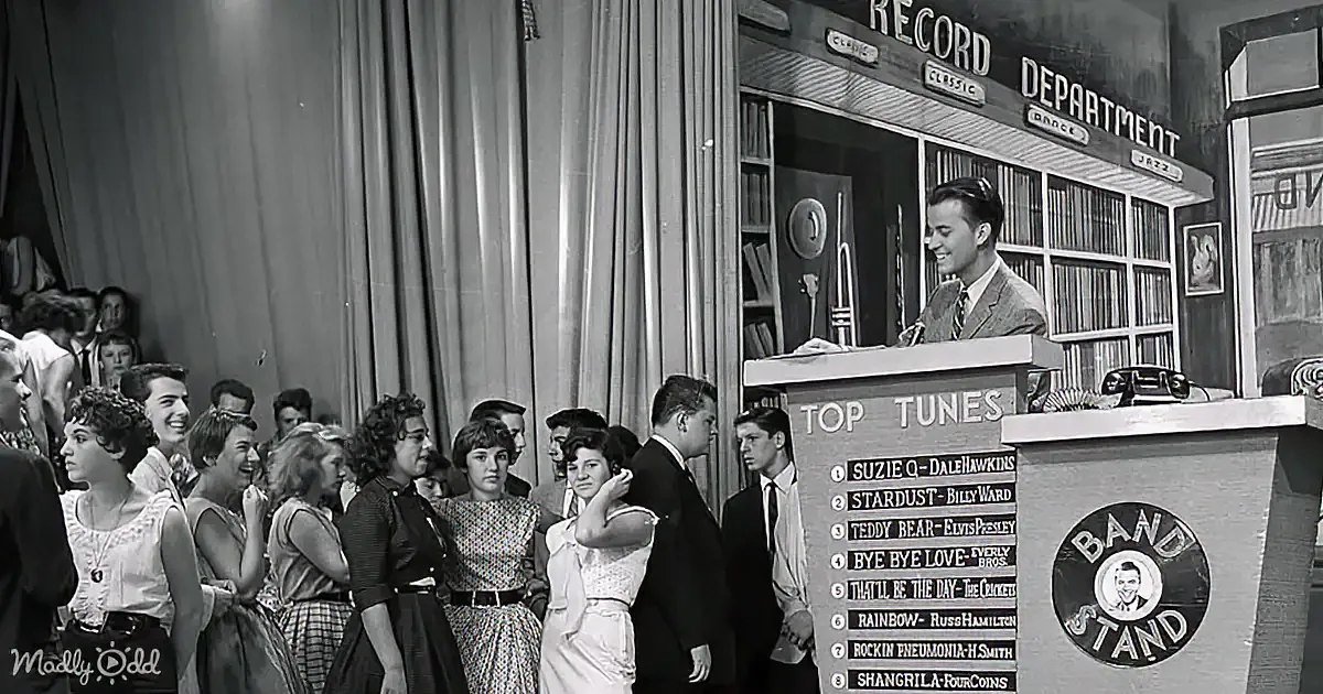 A man stands at a podium with a list of "Top Tunes" in a lively setting, surrounded by a crowd of people.