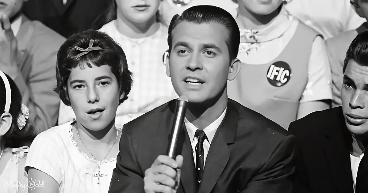 A man in a suit holds a microphone, surrounded by a group of people in a black-and-white setting.
