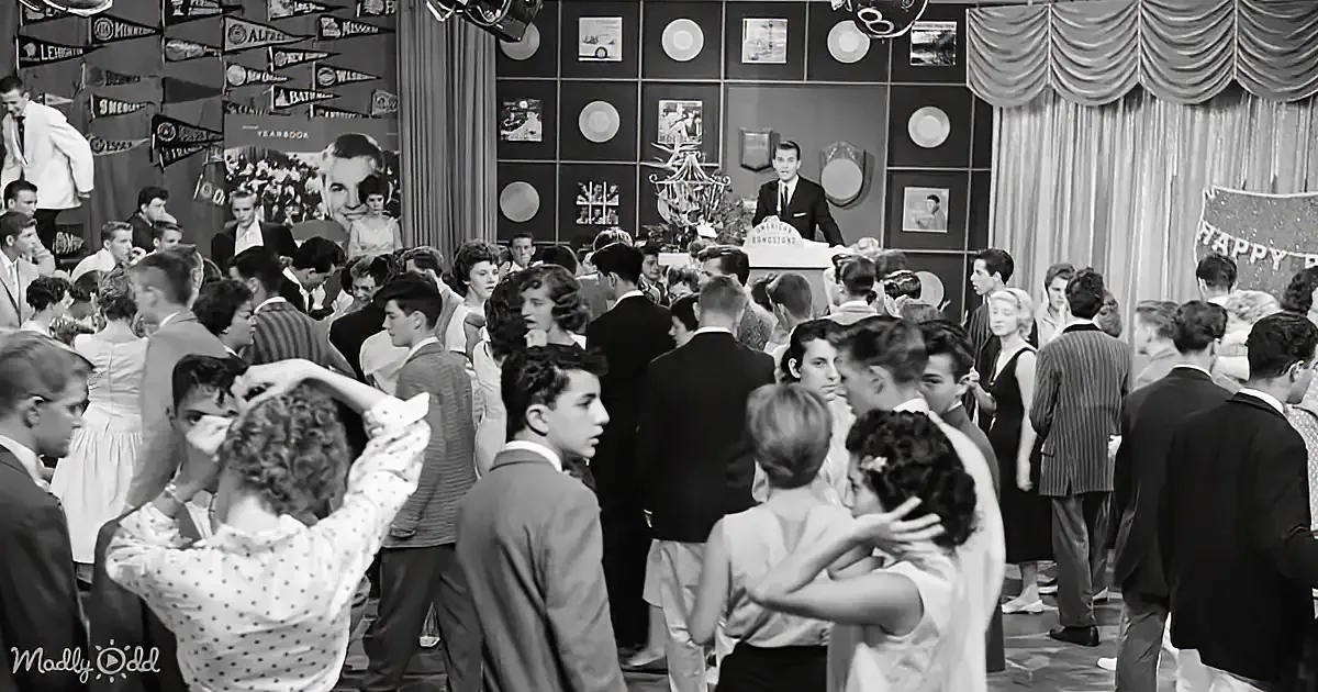 A man in a suit stands at a podium addressing a crowded dance floor.
