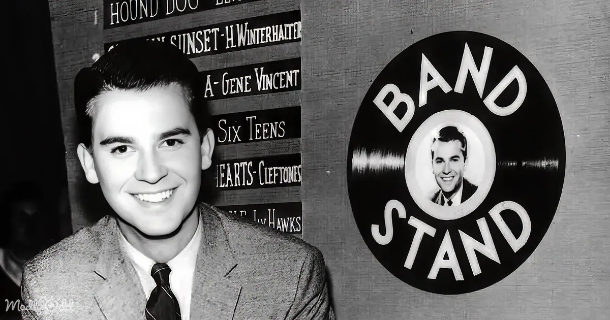 A smiling man in a suit stands beside a "Bandstand" sign with Gene Vincent's image, in a vintage setting.