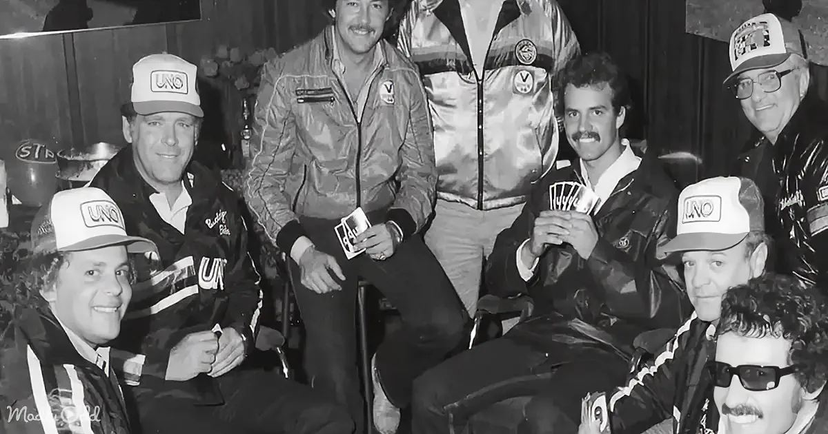 A group of men in racing jackets and caps pose together, smiling, holding cards.