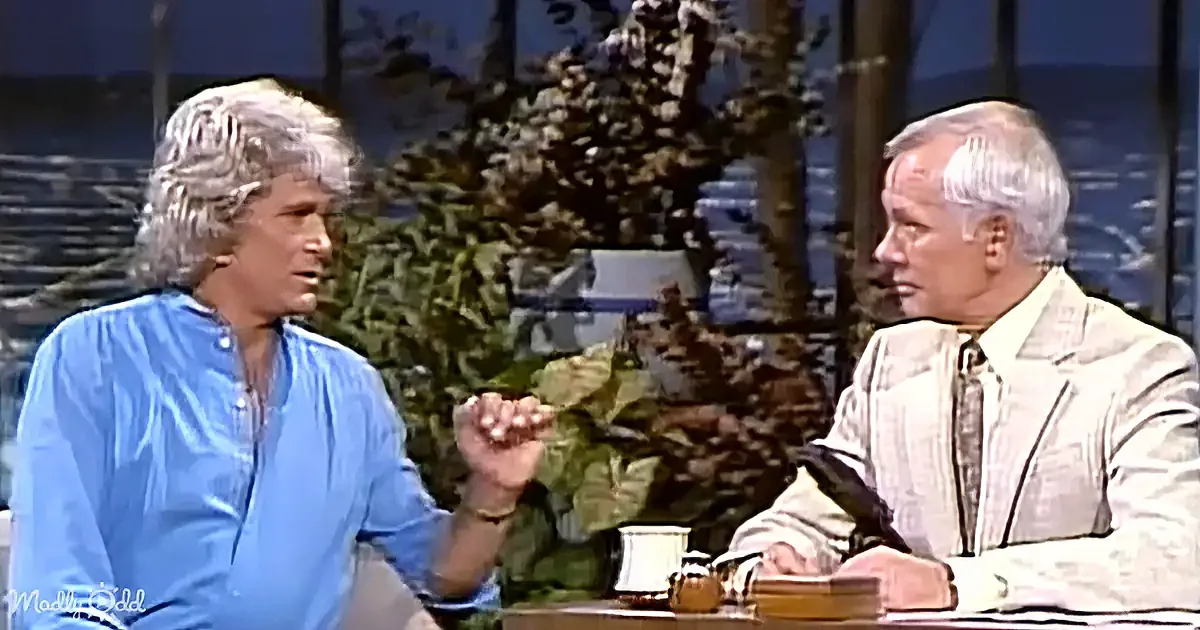 A man in a blue shirt gestures while speaking to another man in a white suit, both seated at a table with a mug.