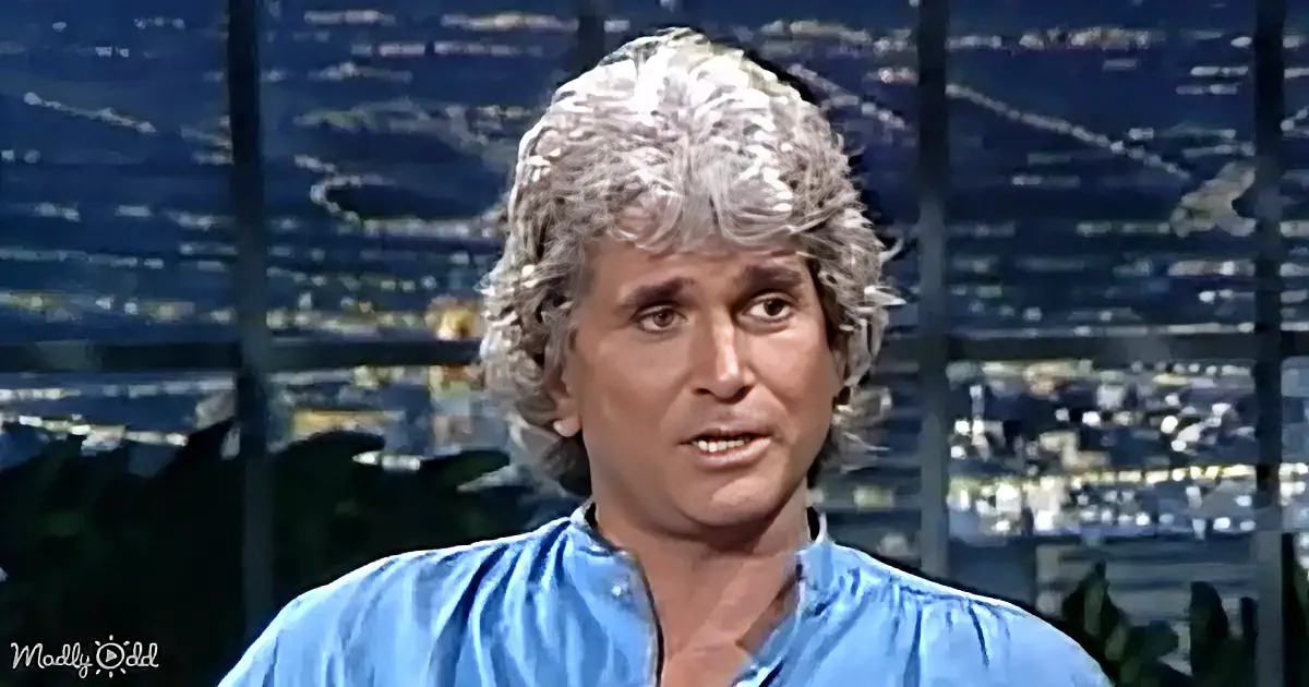A person with curly gray hair, wearing a blue shirt, is speaking on a talk show set with a cityscape background.