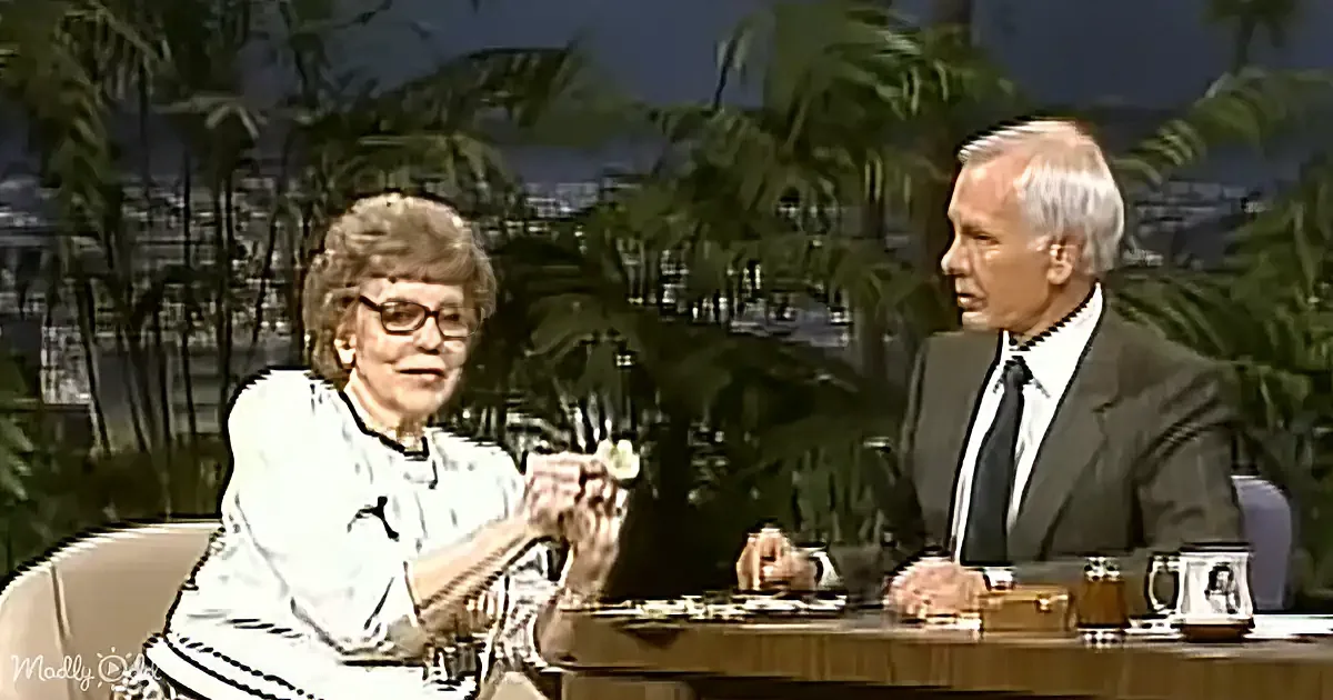 An older woman in glasses holds a small object, seated at a table with a man in a suit, both in a studio setting with...