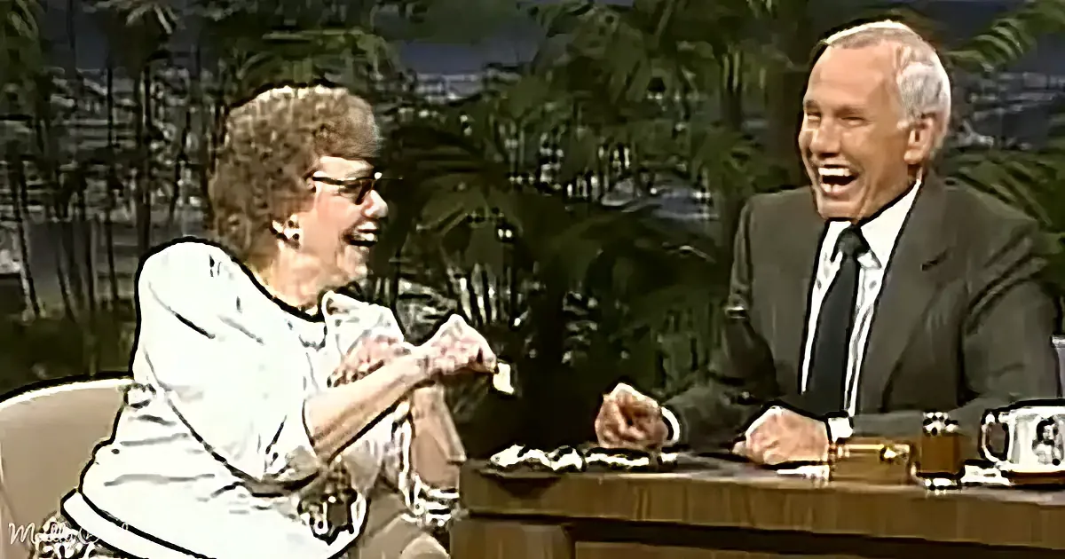 An older woman in a white blouse and glasses is laughing, holding a small object, while a man in a suit and tie sits...