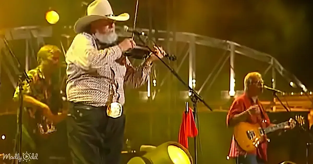 A man in a cowboy hat plays a fiddle on stage, surrounded by other musicians.