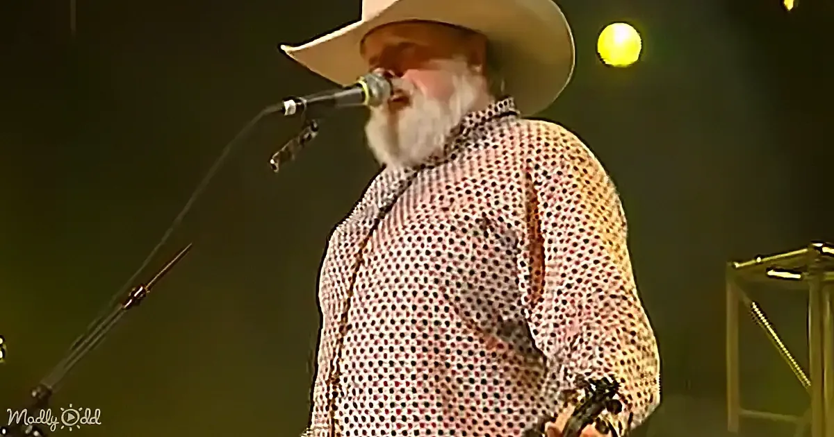 A man with a white beard and cowboy hat sings into a microphone on stage.
