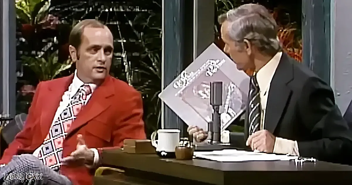 A man in a red suit is seated at a desk, gesturing while speaking to another man in a suit.