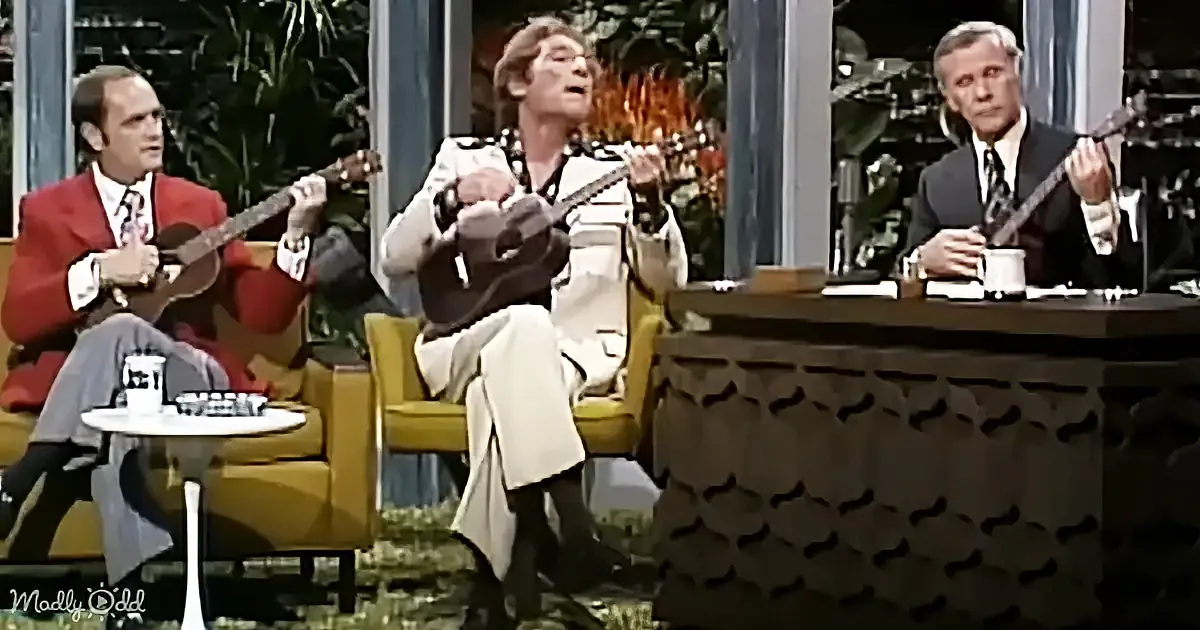 Three men play ukuleles on a TV show set with a host at a desk.
