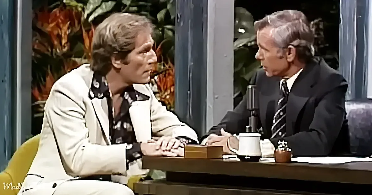Two men in suits sit at a table, one in white, the other in dark, discussing in a studio setting.