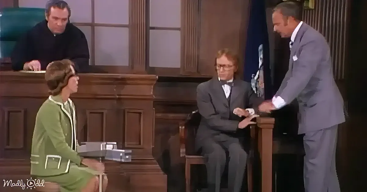 A man in a suit stands at a podium, addressing a seated audience in a courtroom setting.