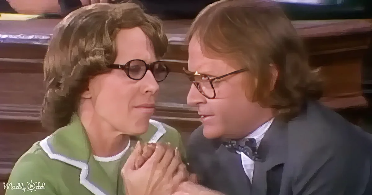 A man and woman are seated closely, the man in a suit and glasses, the woman in a green sweater, both appearing engaged...