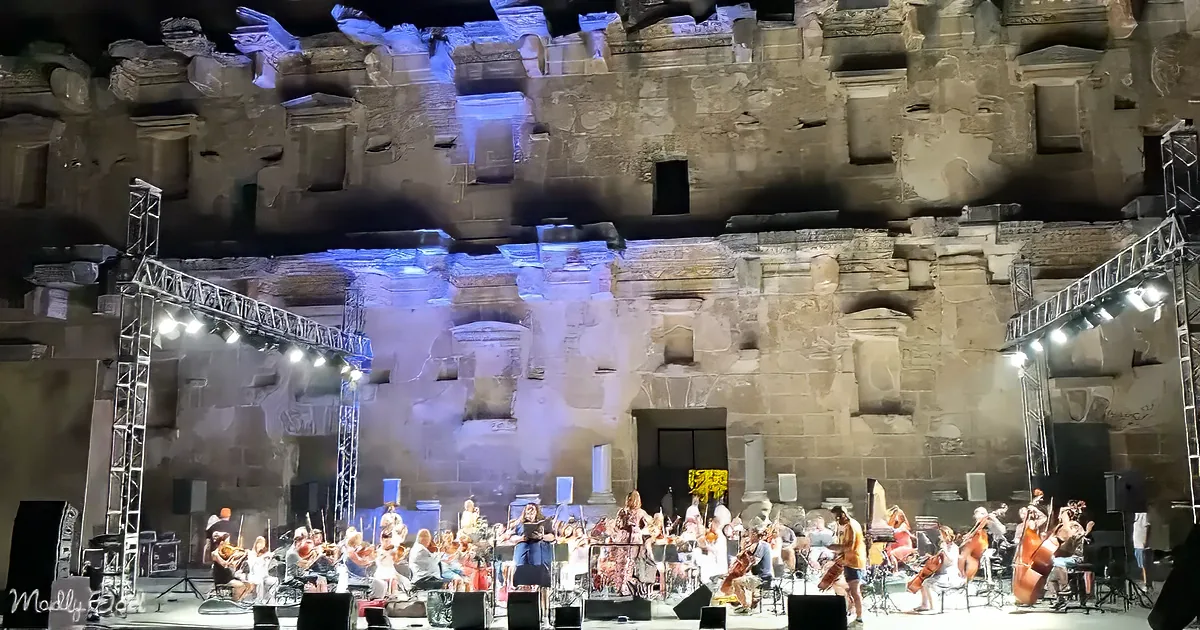 An orchestra performs under ancient stone architecture with blue lighting and stage equipment.