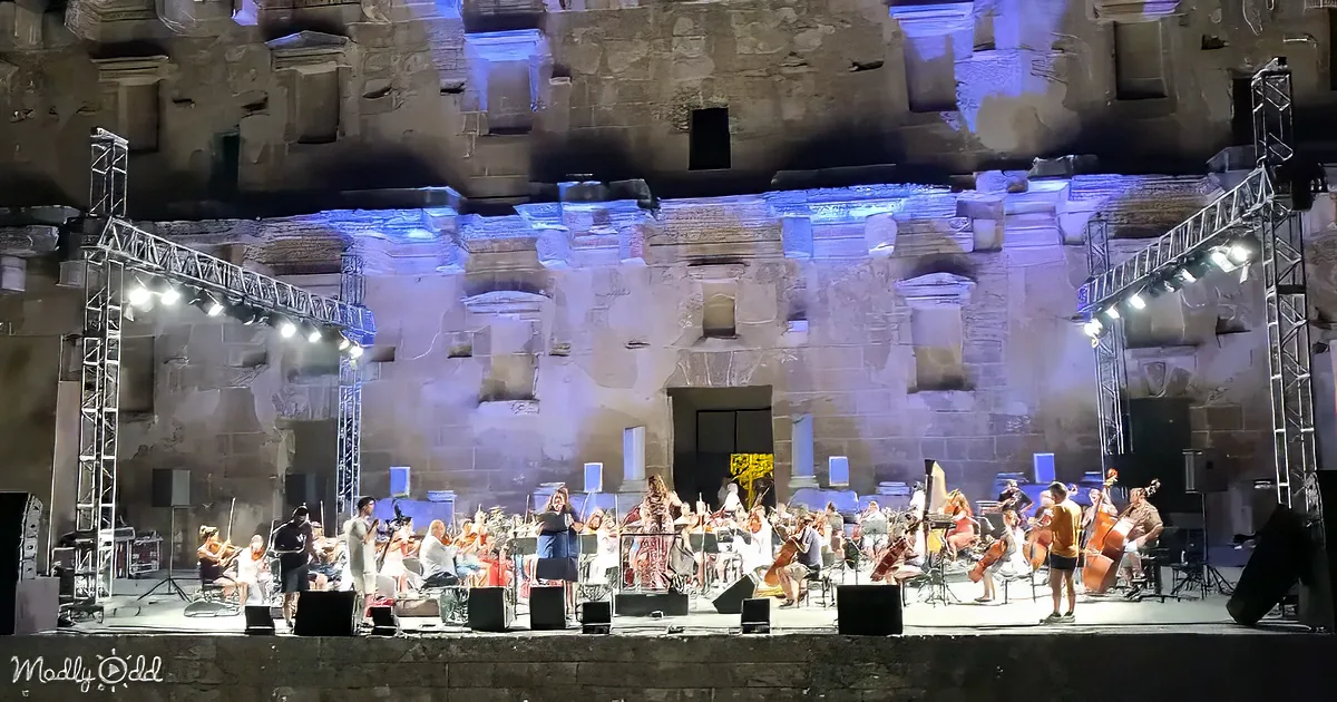 An orchestra performs on stage in front of a historic stone structure illuminated by blue lights.