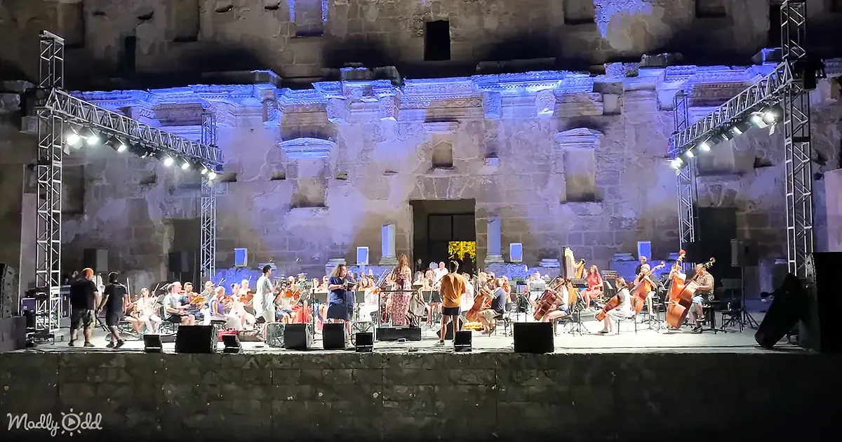 An orchestra performs on stage in front of ancient stone ruins under bright lights.