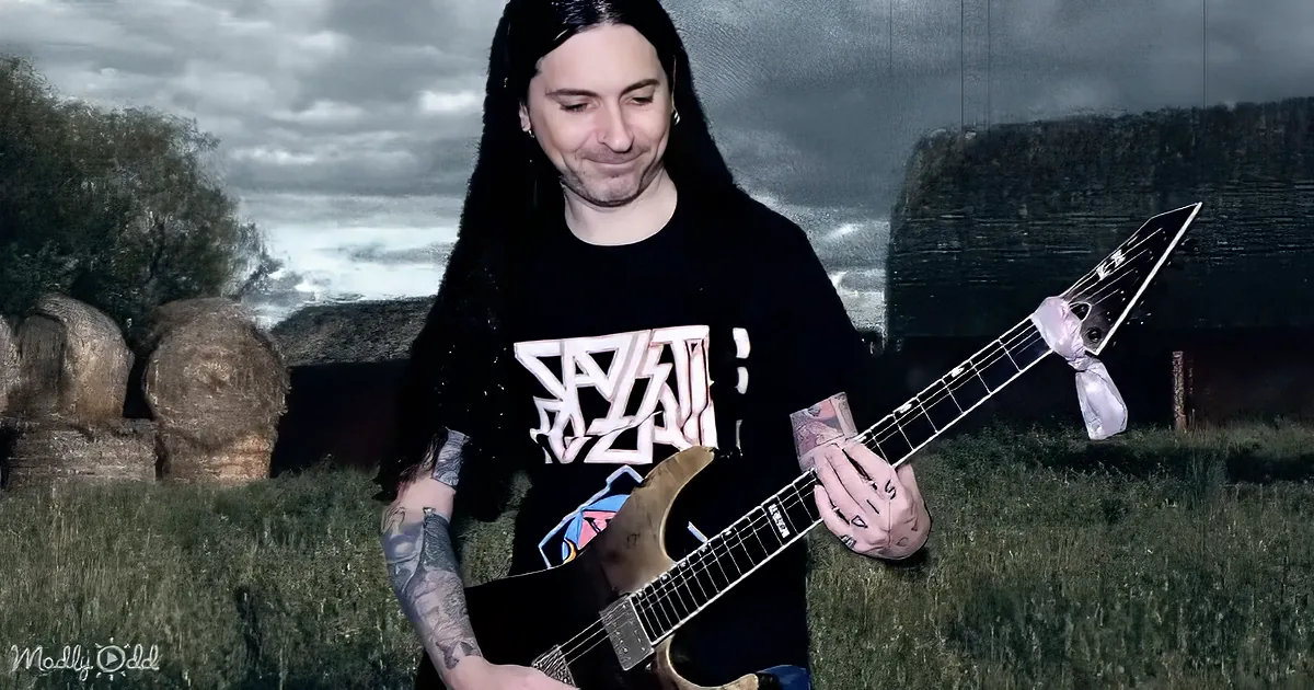 A person with long dark hair and tattoos plays a guitar in a field with hay bales and a cloudy sky.