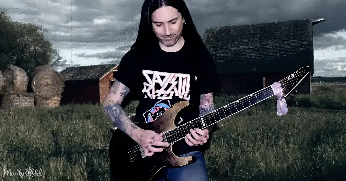 A person with tattoos plays a guitar in a field with a barn and hay bales in the background.