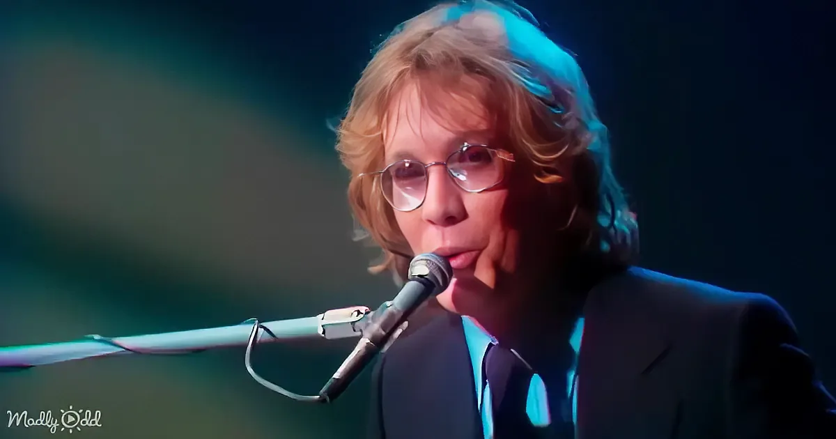 A person with curly hair and glasses sings into a microphone, wearing a dark suit and tie.