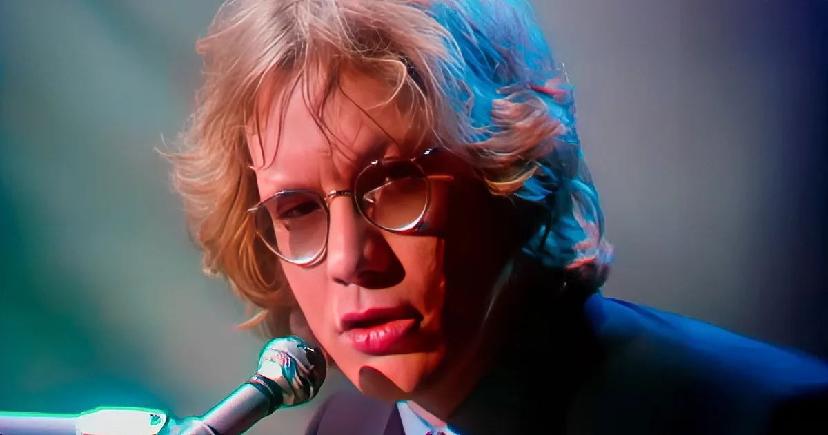 A person with long, wavy hair and glasses is singing into a microphone, with a blurred background and colorful lighting.