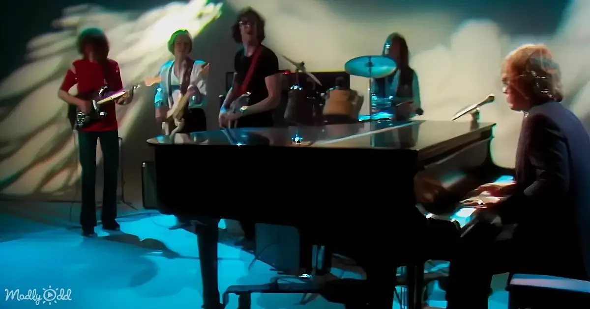 A person plays piano on stage with a band, including guitarists and a drummer, under dramatic lighting.
