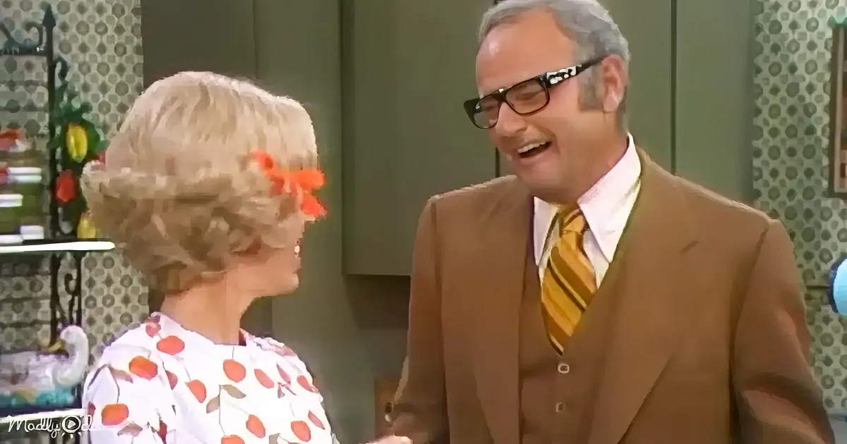 A man in a brown suit and glasses laughs while standing next to a woman with blonde hair and a floral dress.