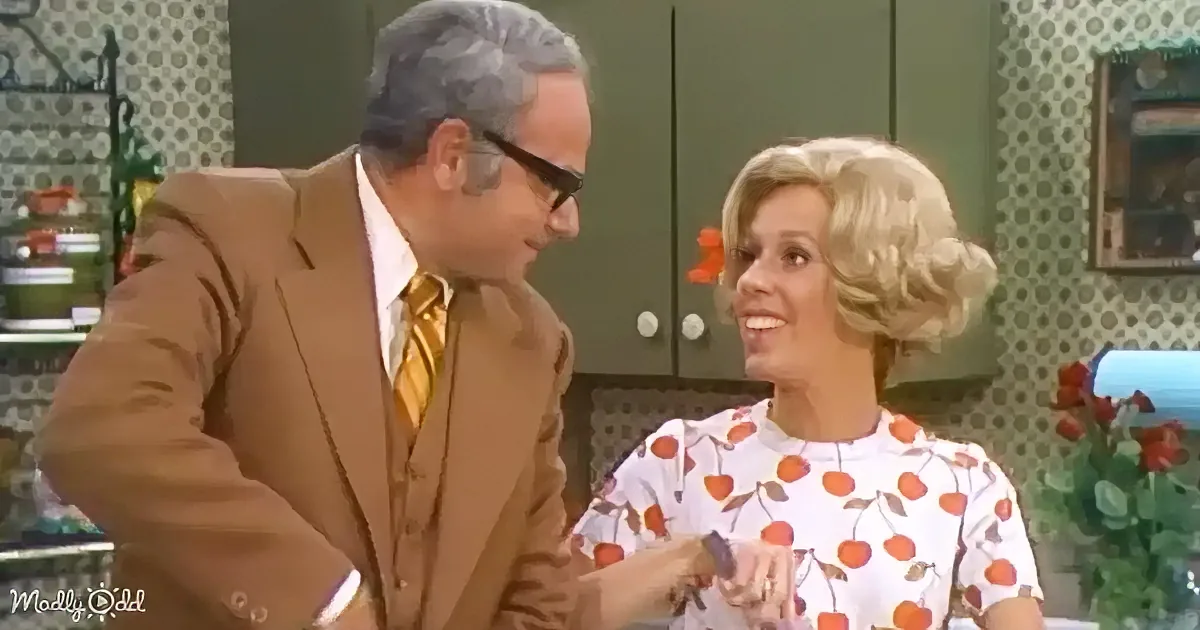 A man in a brown suit and a woman in a floral dress are having a conversation in a kitchen setting.