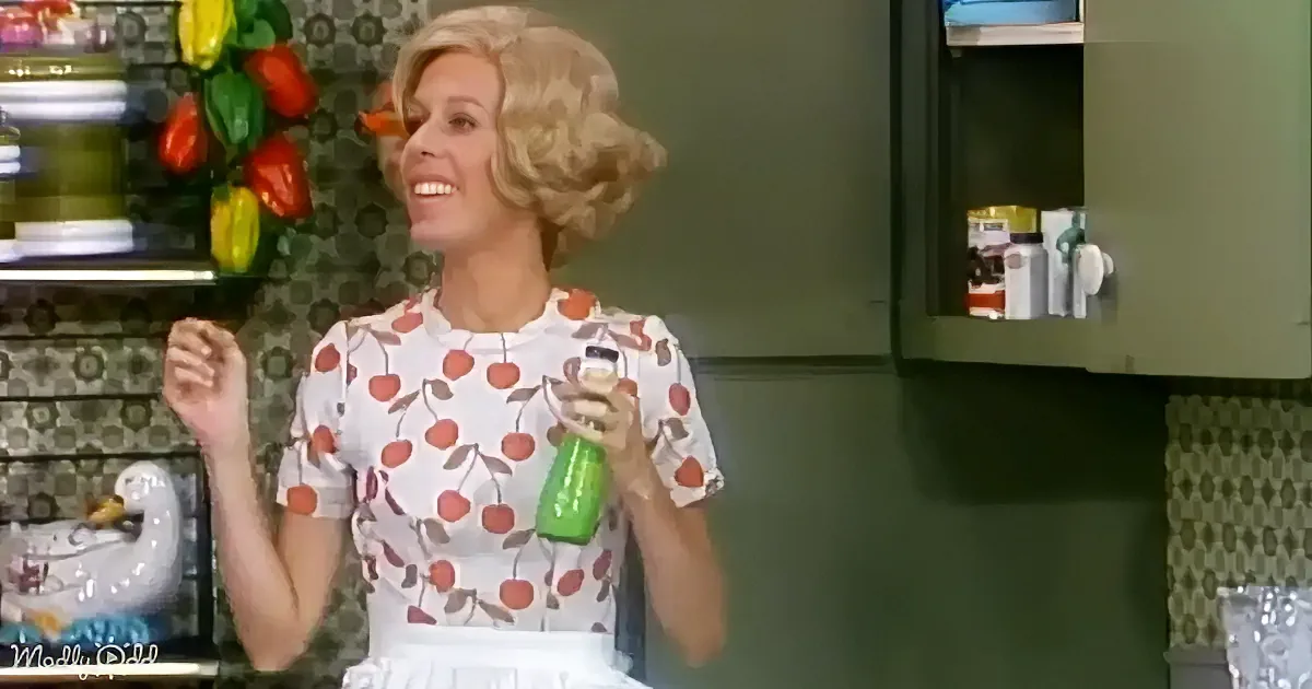 A woman in a floral dress holds a green bottle, smiling in a kitchen with patterned wallpaper and cabinets.