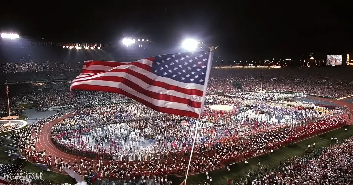 A large American flag waves prominently in a stadium filled with spectators during a nighttime event.