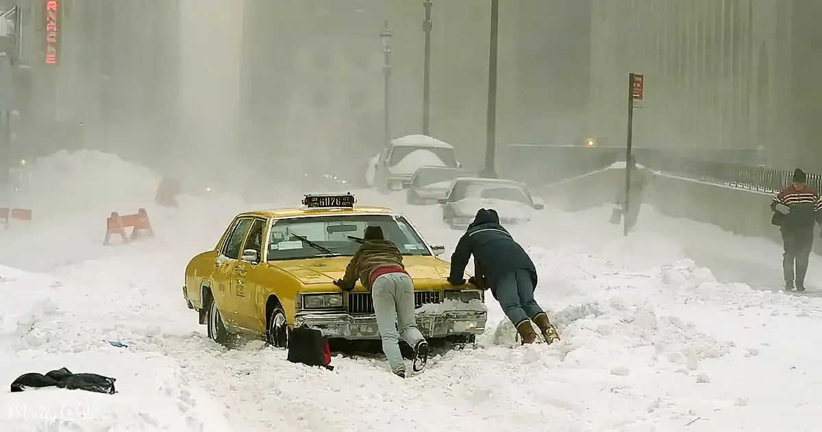 Two people push a yellow taxi stuck in deep snow on a city street.