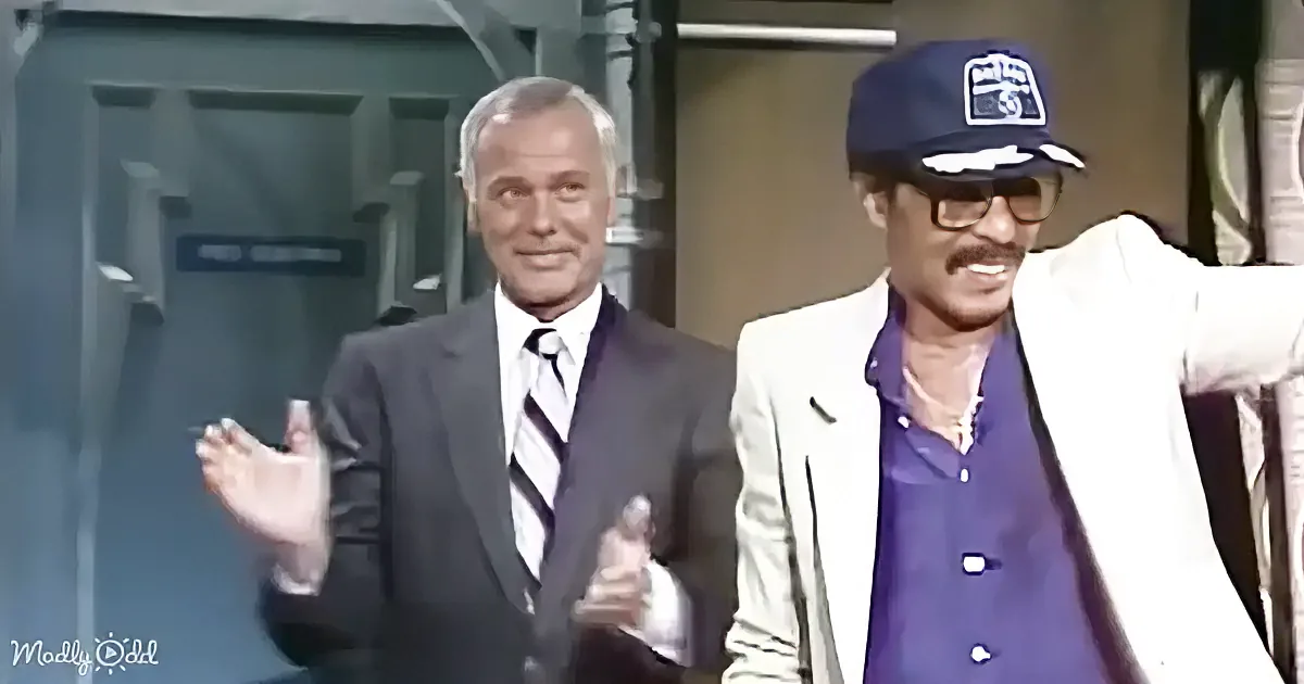 A man in a suit gestures while another in a white blazer and cap leans against a railing.