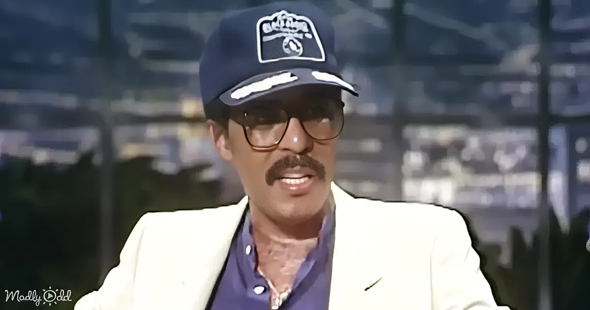 A man wearing a white blazer, blue shirt, and a dark cap with a logo is smiling.