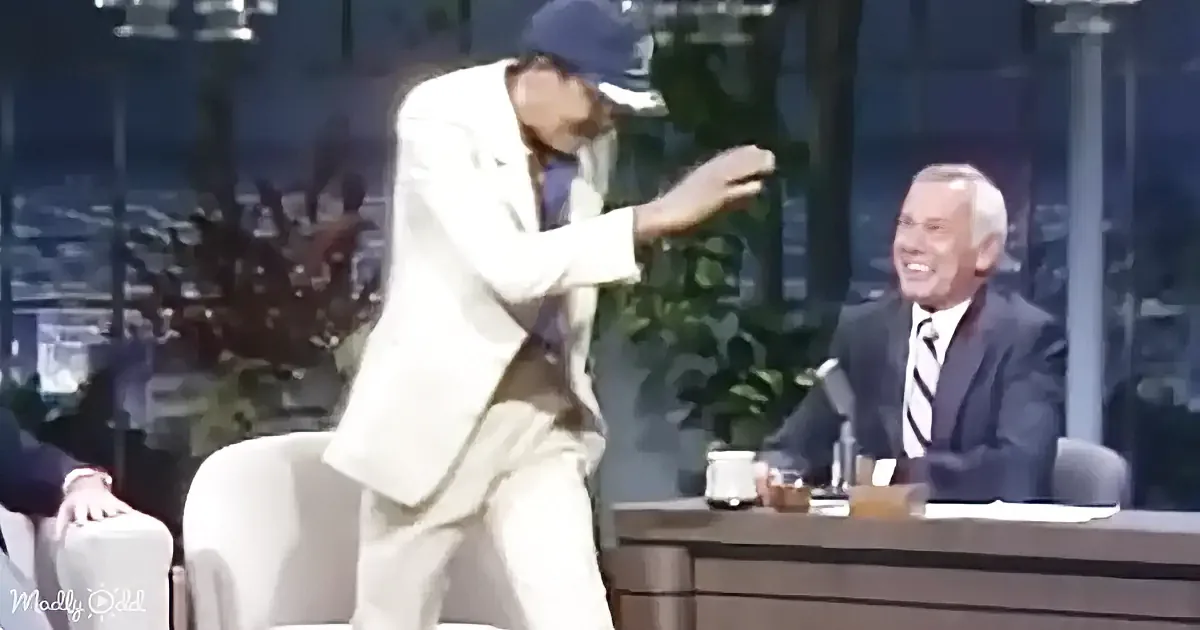 A man in a white suit and cap dances on a talk show set, while a host in a suit sits behind a desk.