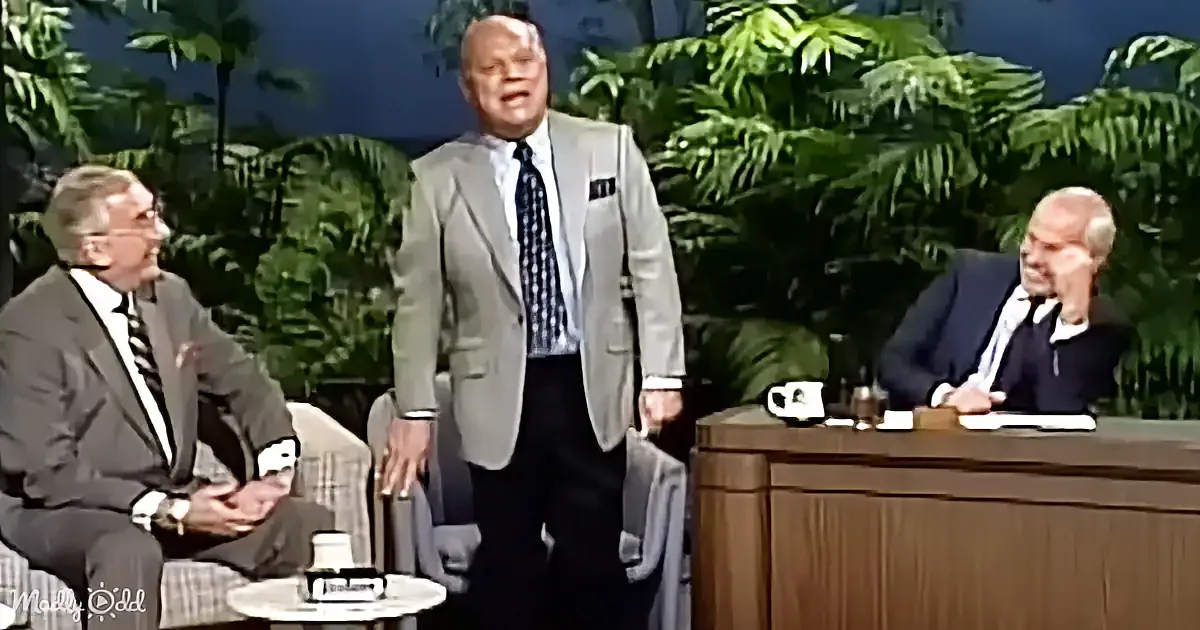 A man in a suit stands, gesturing, while two seated men observe in a studio setting with tropical plants.