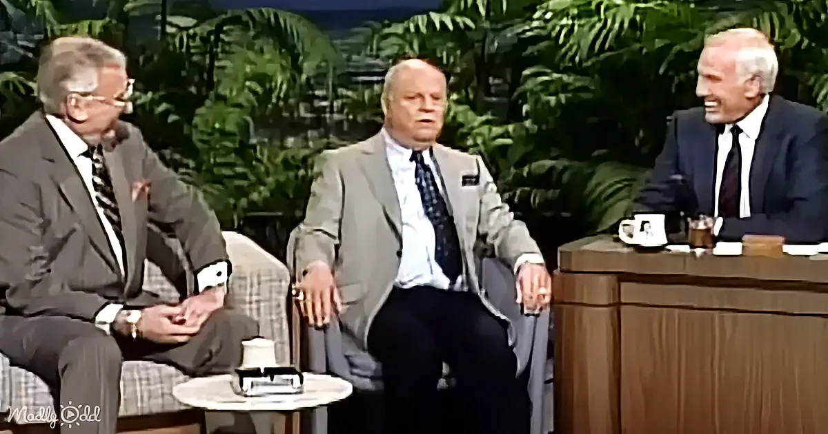Three men in suits sit on a talk show set with a host, surrounded by greenery and a coffee table.