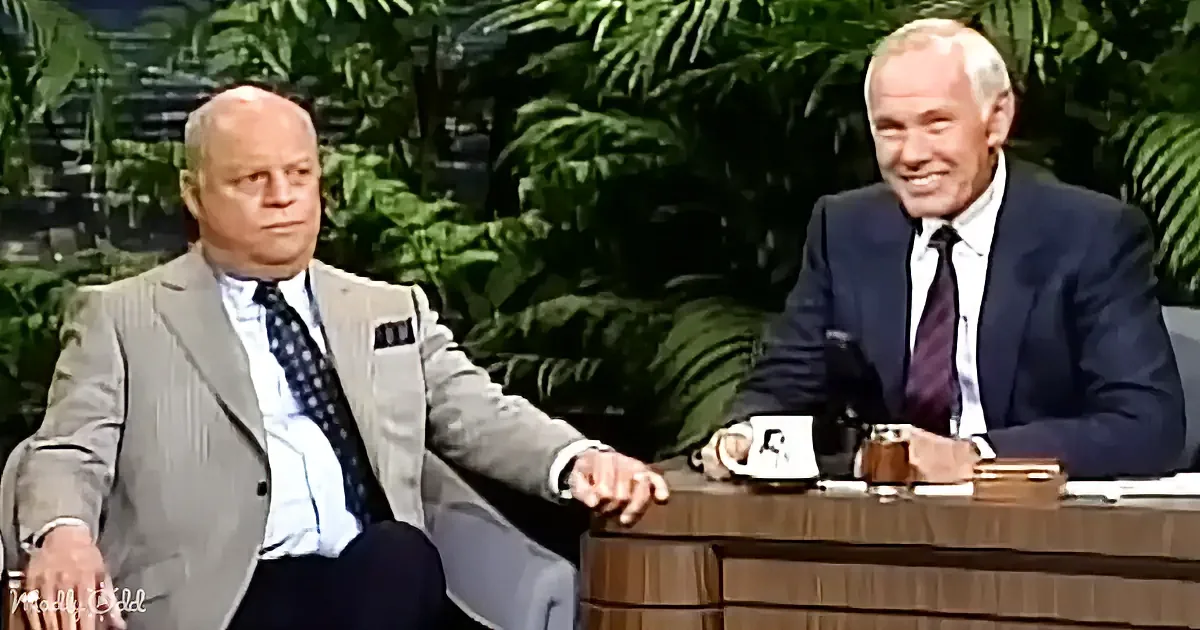 Two men in suits sit at a desk, one gesturing, both smiling, with greenery in the background.