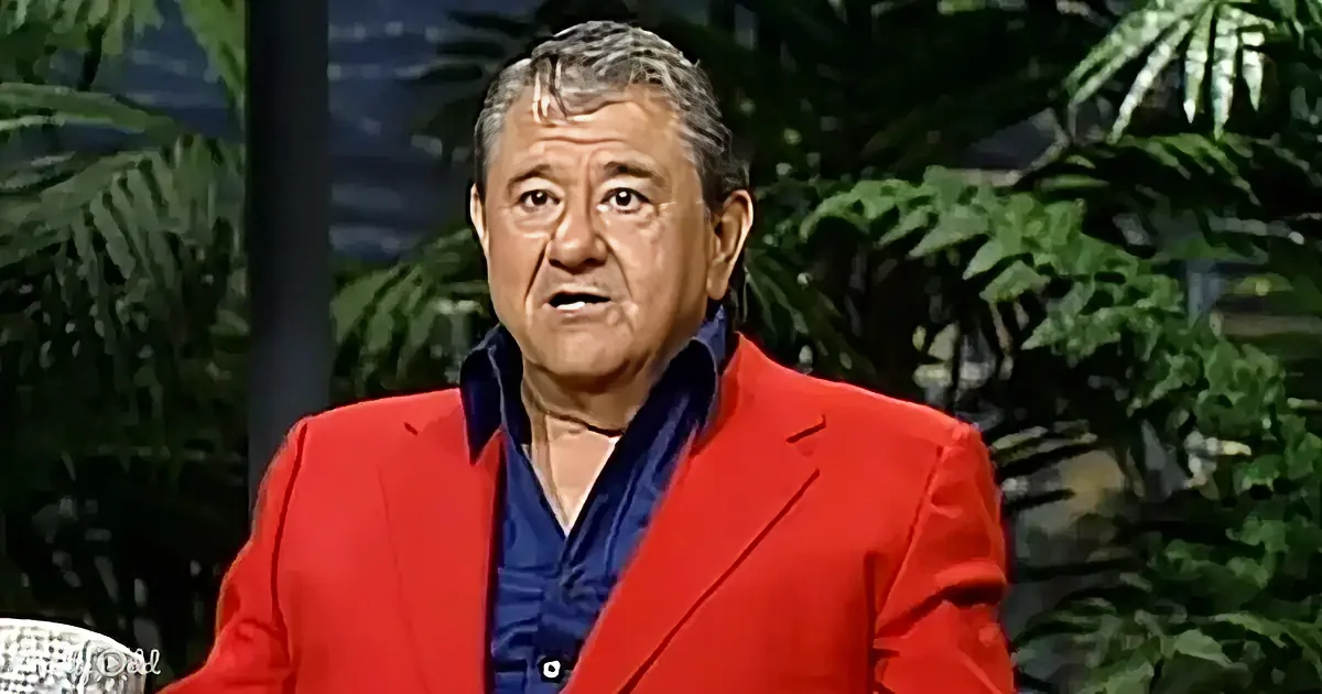 A man in a red suit jacket and blue shirt is speaking, with tropical plants in the background.