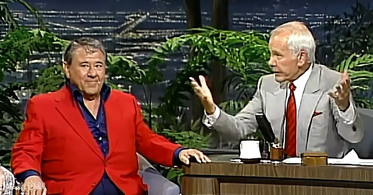 A man in a red suit sits while another in a gray suit gestures animatedly during a talk show.