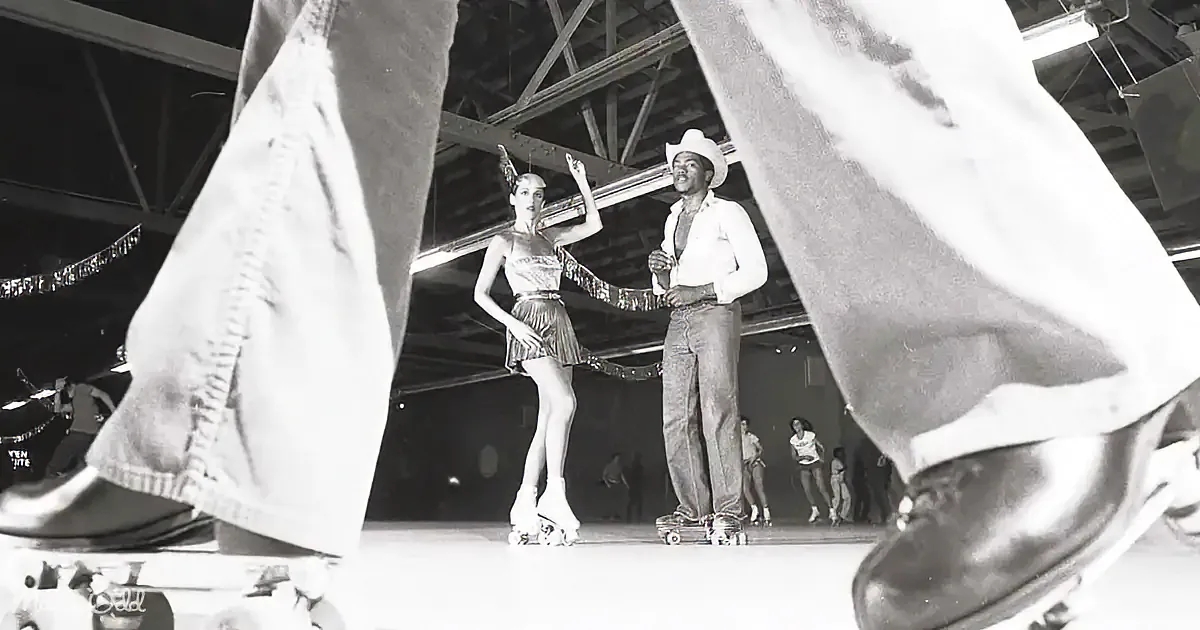 A man and woman roller skate in a vintage setting, surrounded by other skaters and a band.