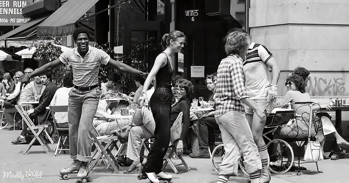 A man and woman roller skate in front of a café, surrounded by seated patrons.