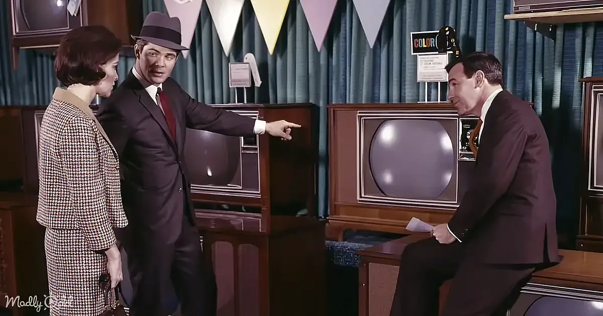A man in a suit points at another man in a suit, both in a room with vintage TVs and decorations.