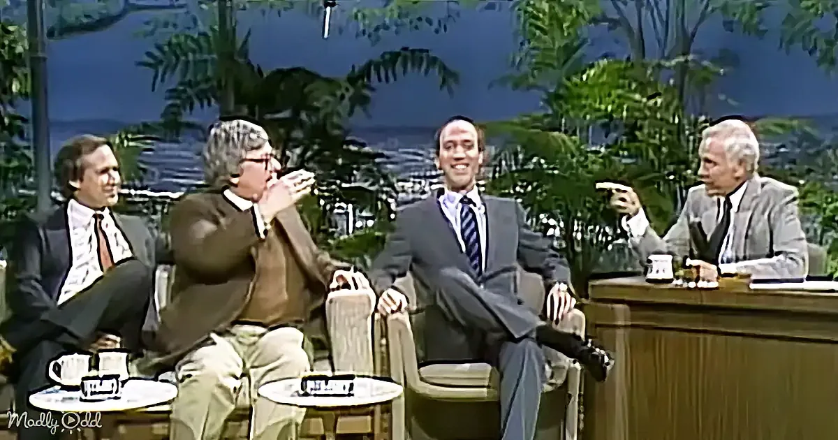 Four men in suits sit on a talk show set with tropical decor, one laughing, another gesturing.