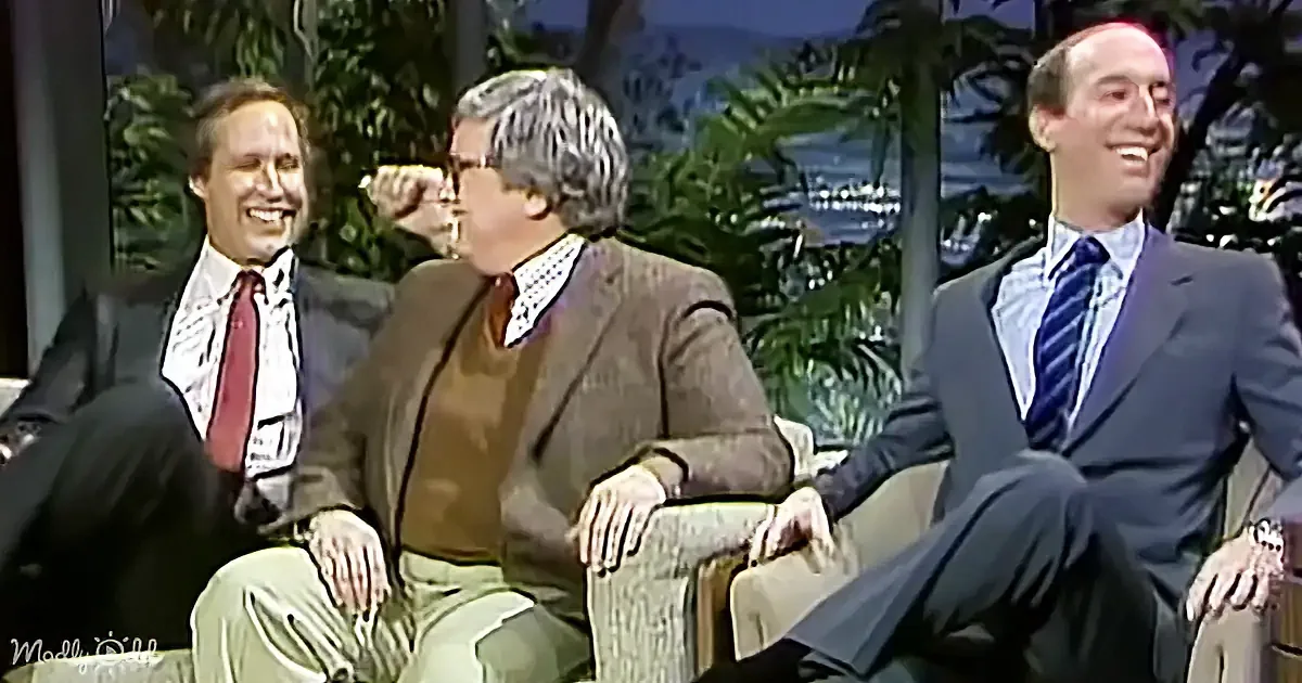 Three men in suits and casual attire are seated, smiling and laughing in a studio setting with tropical decor.