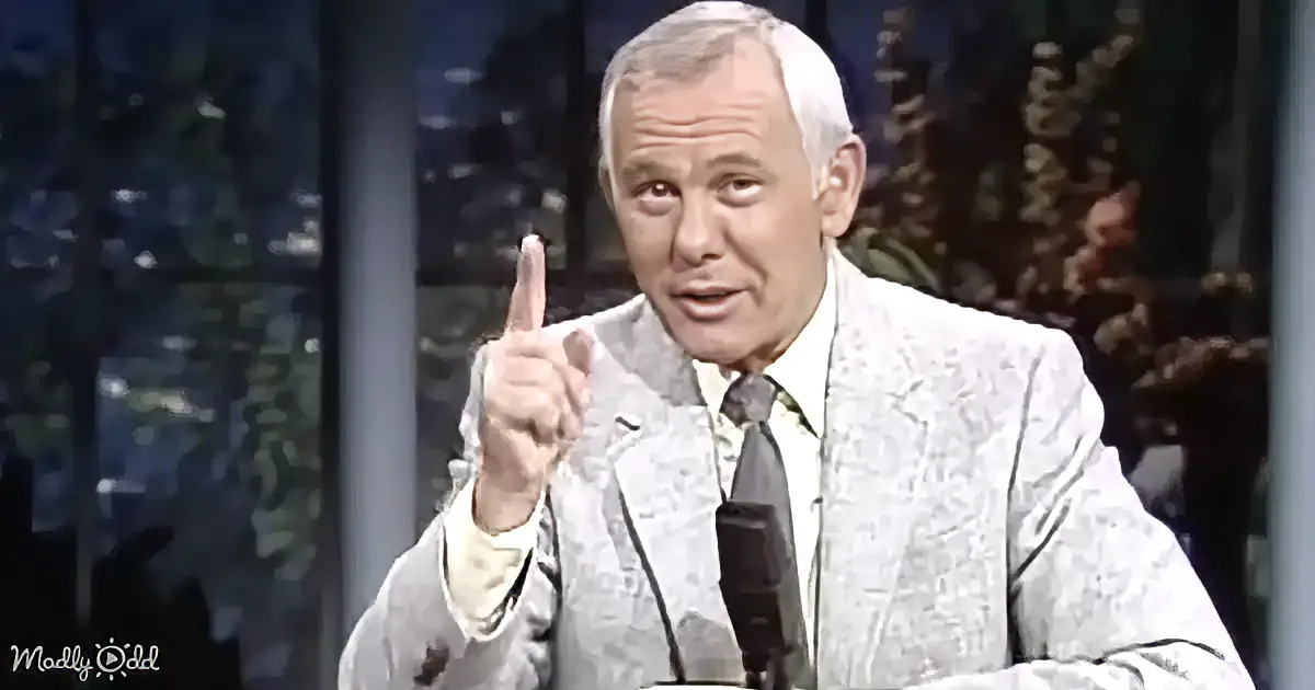 A man in a patterned suit points upwards while speaking on a talk show set.