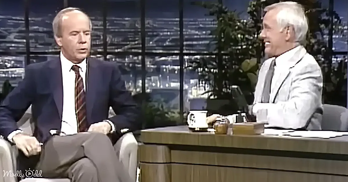 Two men in suits are seated on a talk show set, one in a dark suit and the other in a light suit, with a cityscape backdrop.