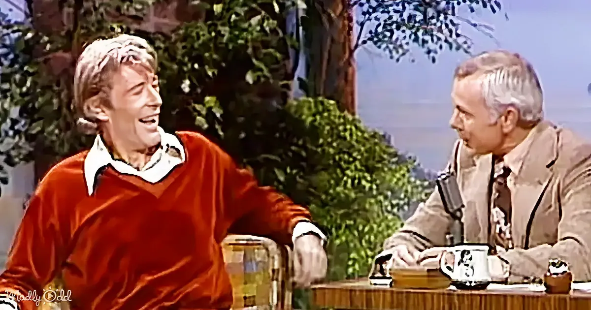 A man in a red sweater laughs while seated at a table with another man in a beige suit, both with microphones, in a...
