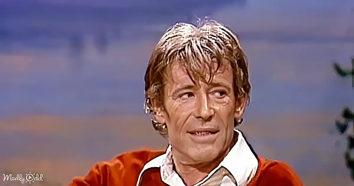 A man with short, light brown hair and a red jacket is looking to the side with a thoughtful expression.