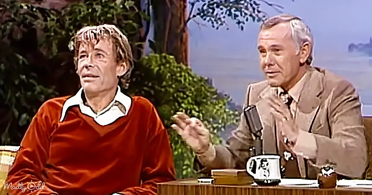 Two men are seated at a table, one in a red sweater gesturing, the other in a suit, both appearing engaged in conversation.